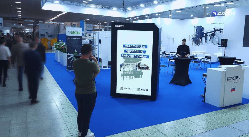 People attend a trade show with blue carpet, exhibition booths, and a large digital sign displaying text in Slovak. One person is taking a photo of the sign, while another stands near a high table.