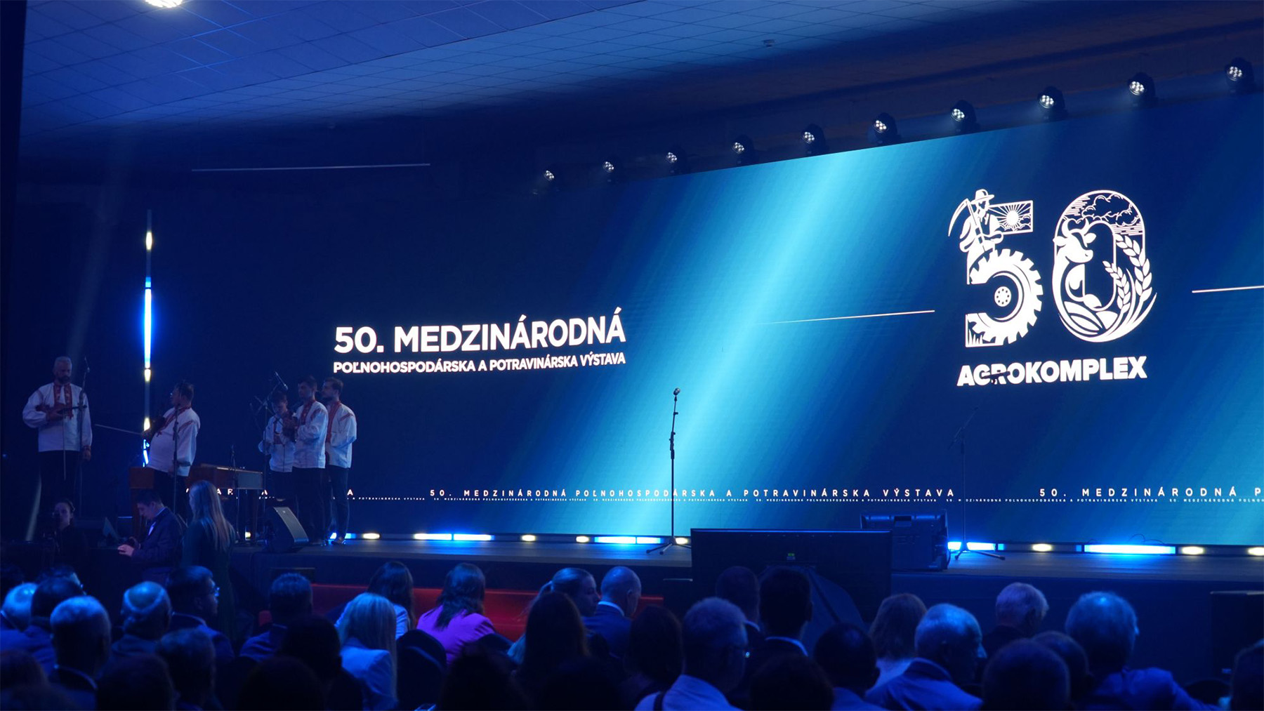 A stage with a large digital screen shows "50. Medzinárodná Agrokoplex" during an event, people onstage, audience seated.