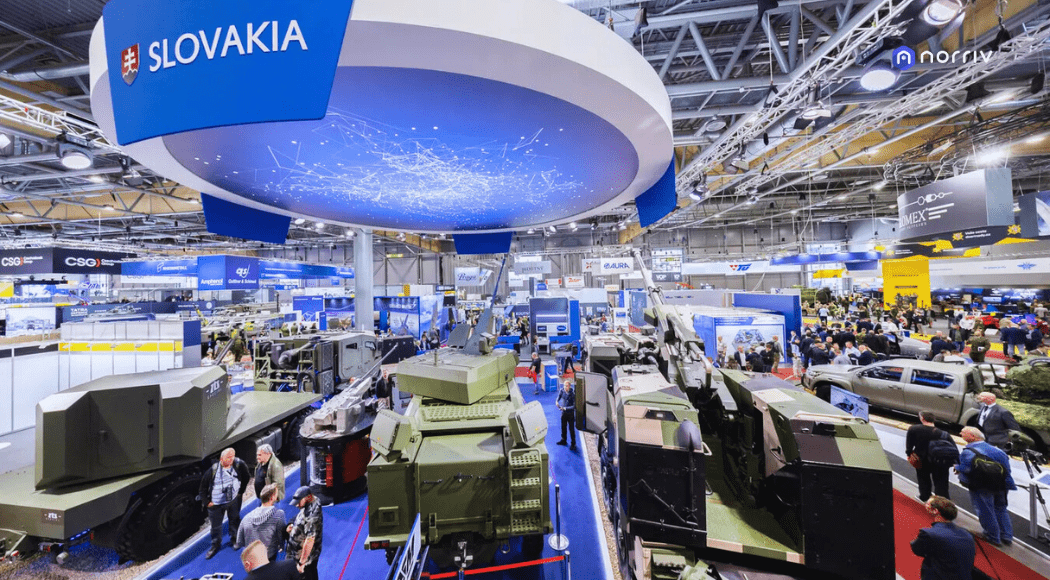 A busy exhibition hall displays military vehicles and technology beneath a large Slovakia sign, with crowds of people walking among various booths and displays at an international defense expo.
