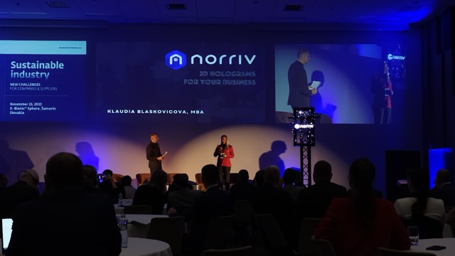 A speaker and a host stand on stage with a projected Norriv 3D Holograms for Your Business logo behind them. Audience members are seated in a dimly lit conference room. A Sustainable Industry event slide is visible on the left.