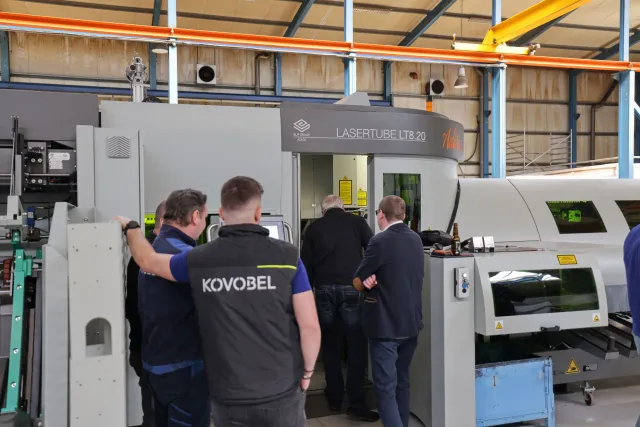 Four men stand around a large industrial machine labeled LASERTUBE LT8.20 in a factory setting, with one man appearing to operate or inspect the machine. The workers wear casual and branded workwear.