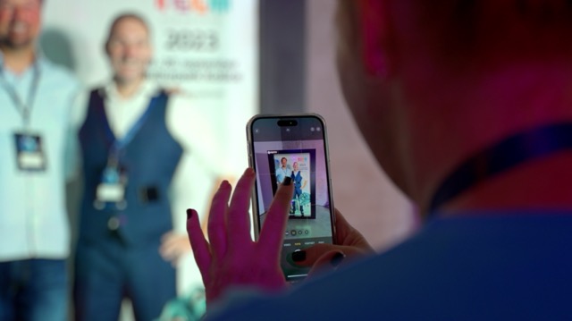 A person takes a photo with a smartphone of two people posing together in front of a colorful event banner. The image focuses on the phone screen, capturing the two subjects.