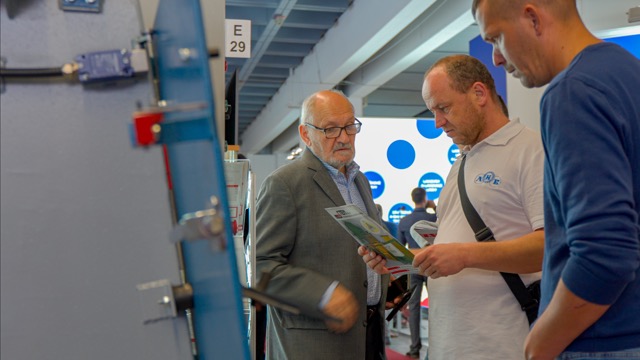 Three men stand indoors at an event or exhibition, one in a suit and two in casual clothes. They examine a brochure together, with machines and informational displays visible in the background.