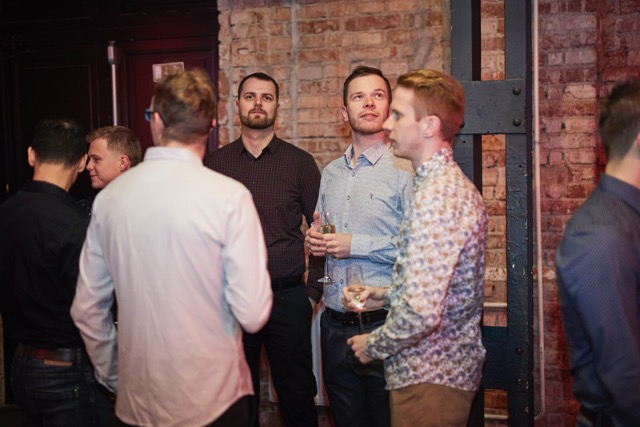 A group of men in casual shirts stand and talk at an indoor event, holding drinks. They are gathered near a brick wall and metal beams, some facing each other and some looking away.