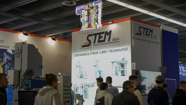 People attend an industrial trade show, gathered around a STEM d.o.o. booth with signage featuring machinery and technology diagrams. Overhead lights illuminate the exhibit area.