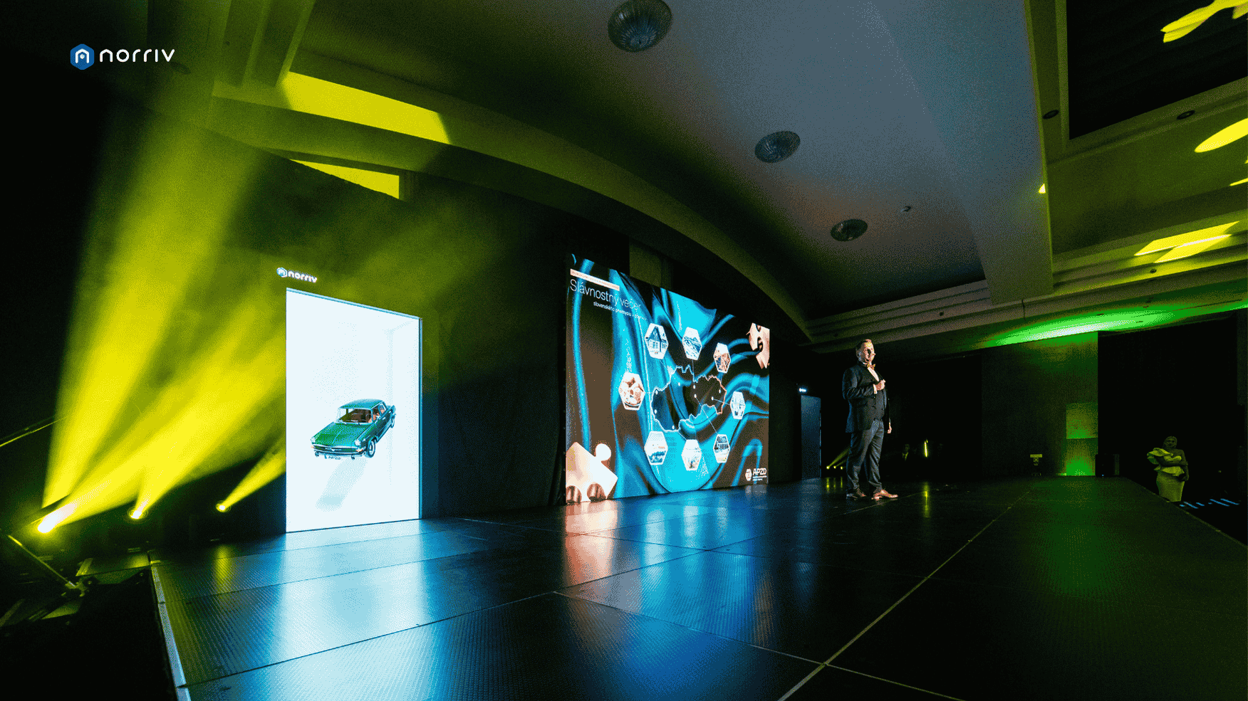 A person stands on a large stage with bright yellow lights and digital screens displaying graphics of technology, cars, and icons. The logo Norriv is visible in the top left corner. The stage is set in a spacious, modern venue.