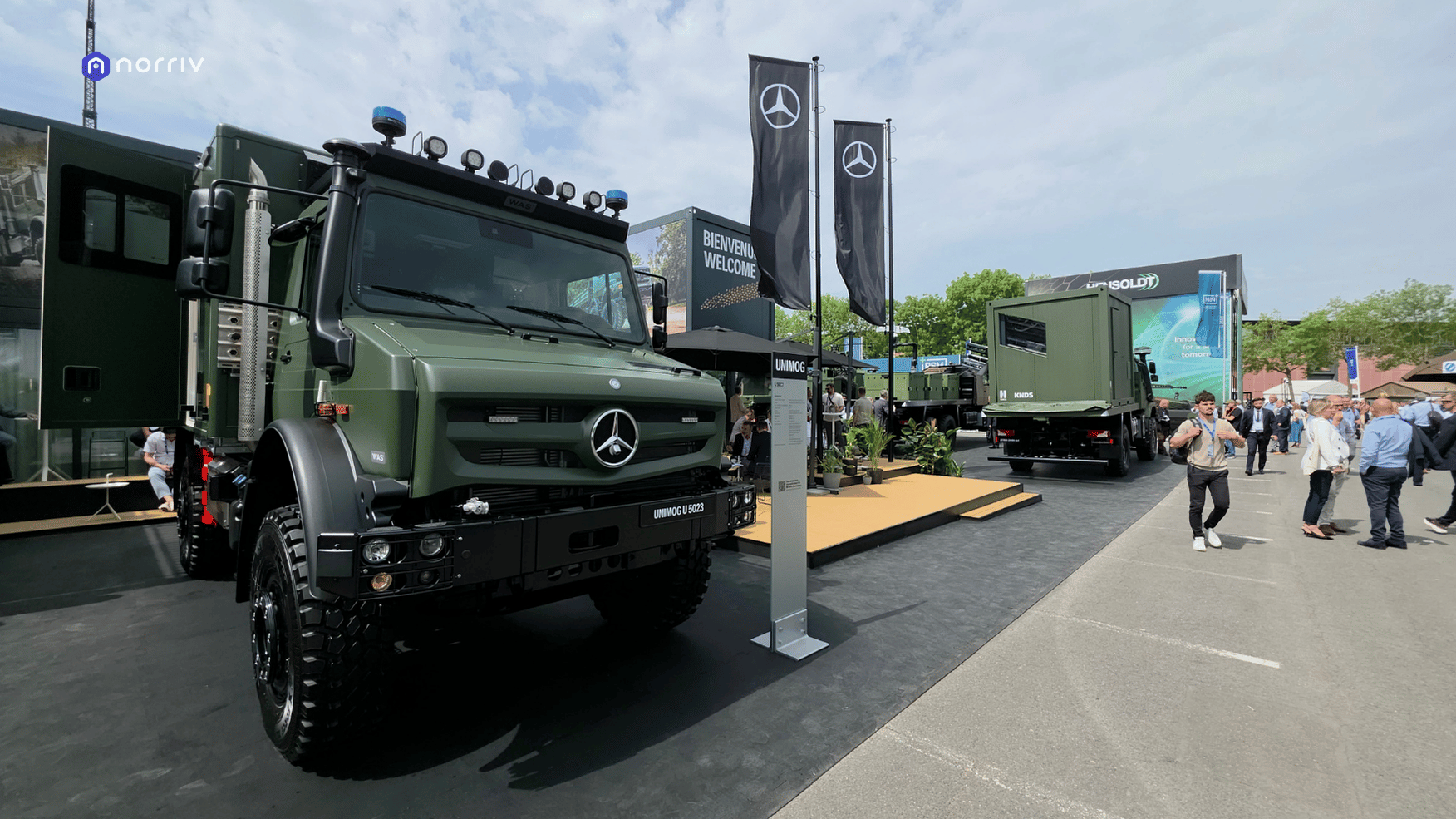 A green Mercedes-Benz Unimog truck and other military vehicles beside visitors in the outdoor display area at Eurosatory 2026.