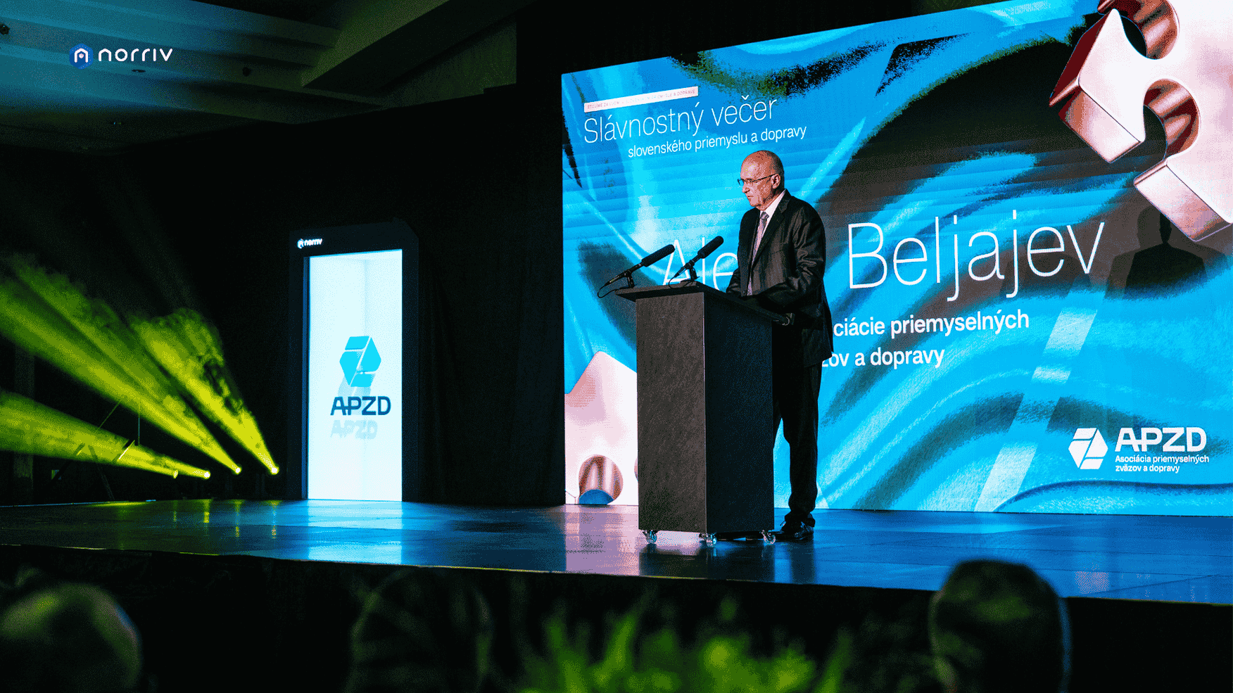 A man in a suit speaks at a podium on a stage with blue lighting, beside a large screen displaying text and a logo for APZD, during a formal event in a modern indoor venue.
