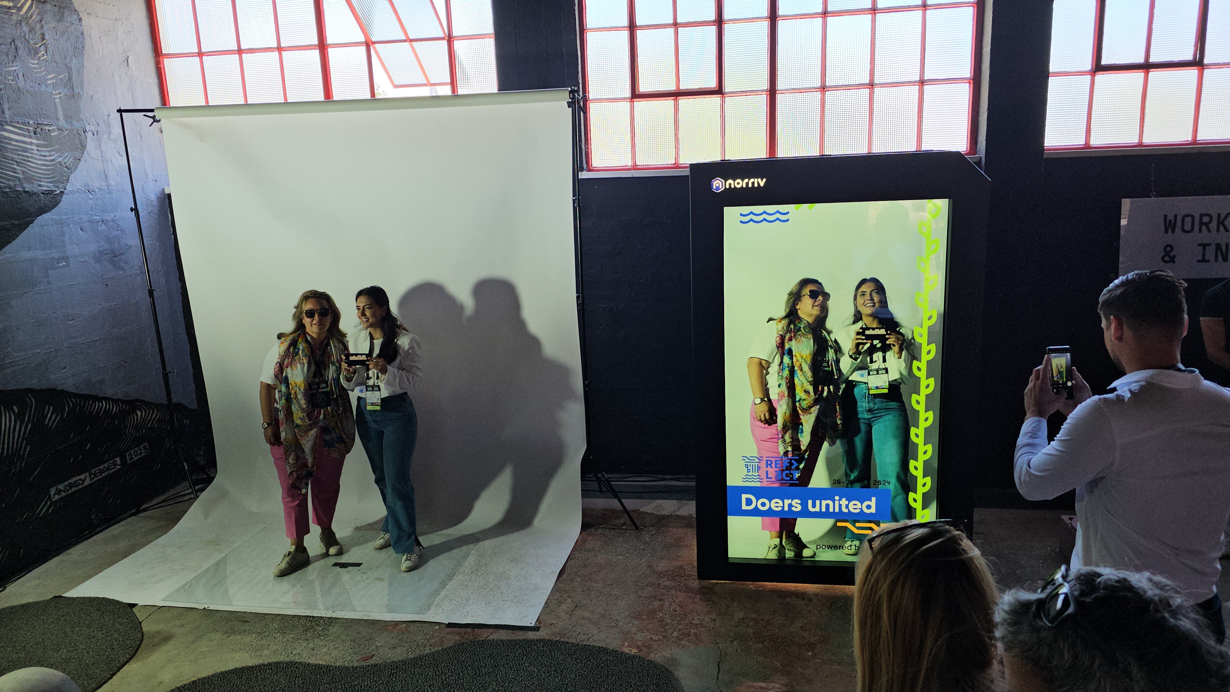 Two women pose for a photo in front of a white backdrop while their image appears on a large digital screen beside them. Several people watch and take photos in an industrial-style room with big windows.
