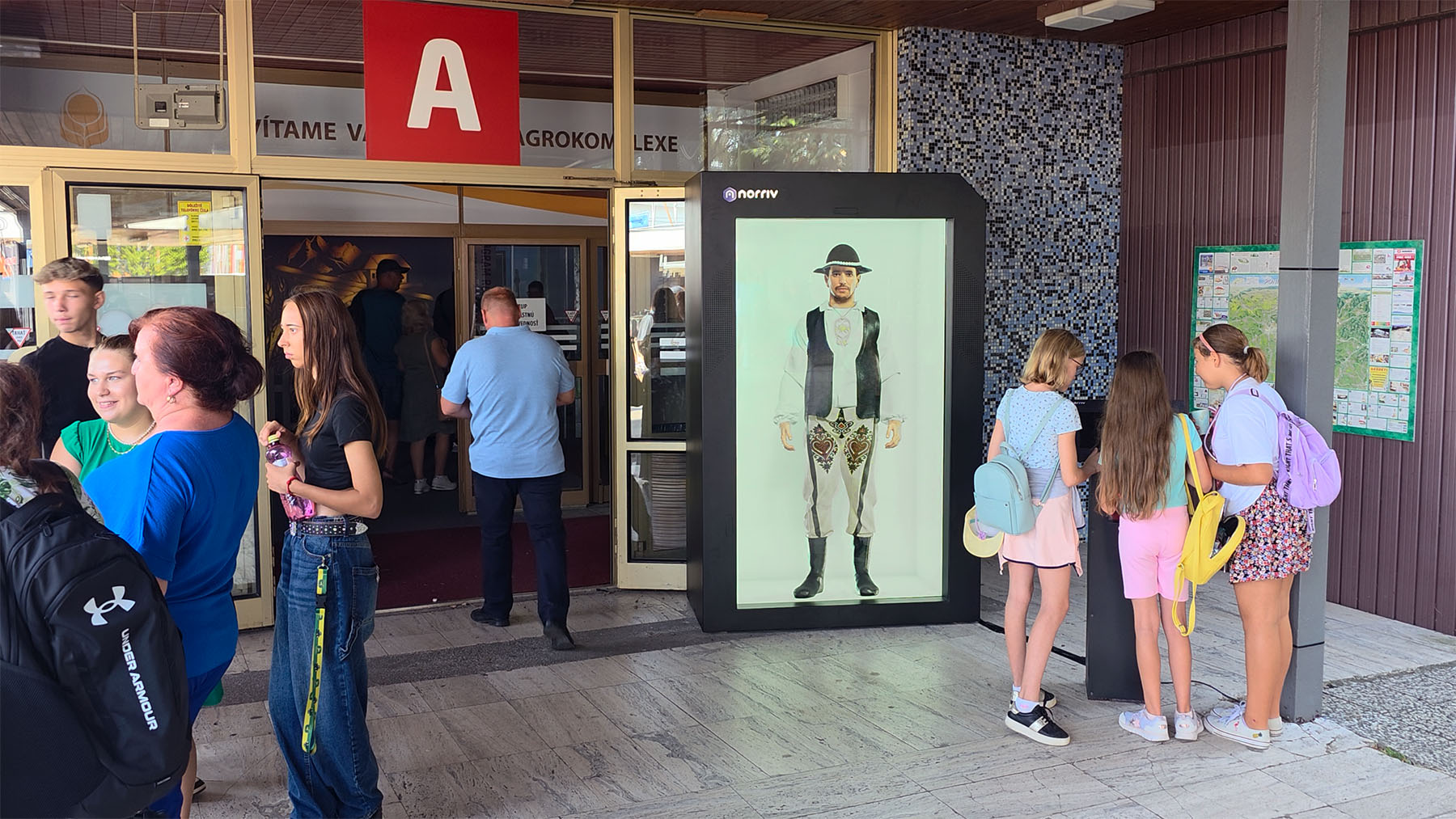 A group stands outside a building entrance; four girls with paddles watch a digital display of a man in traditional attire nearby.