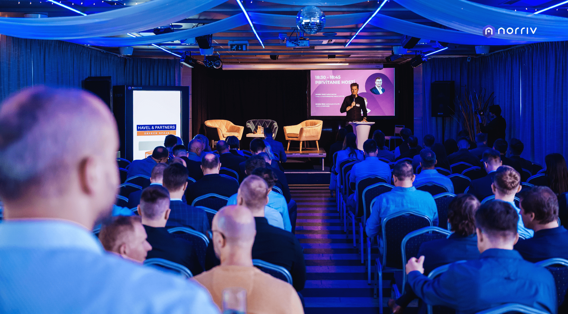 A speaker stands at a podium on a stage addressing an audience seated in rows. The stage is lit with blue lighting, and presentation slides with text and a portrait are displayed behind the speaker.
