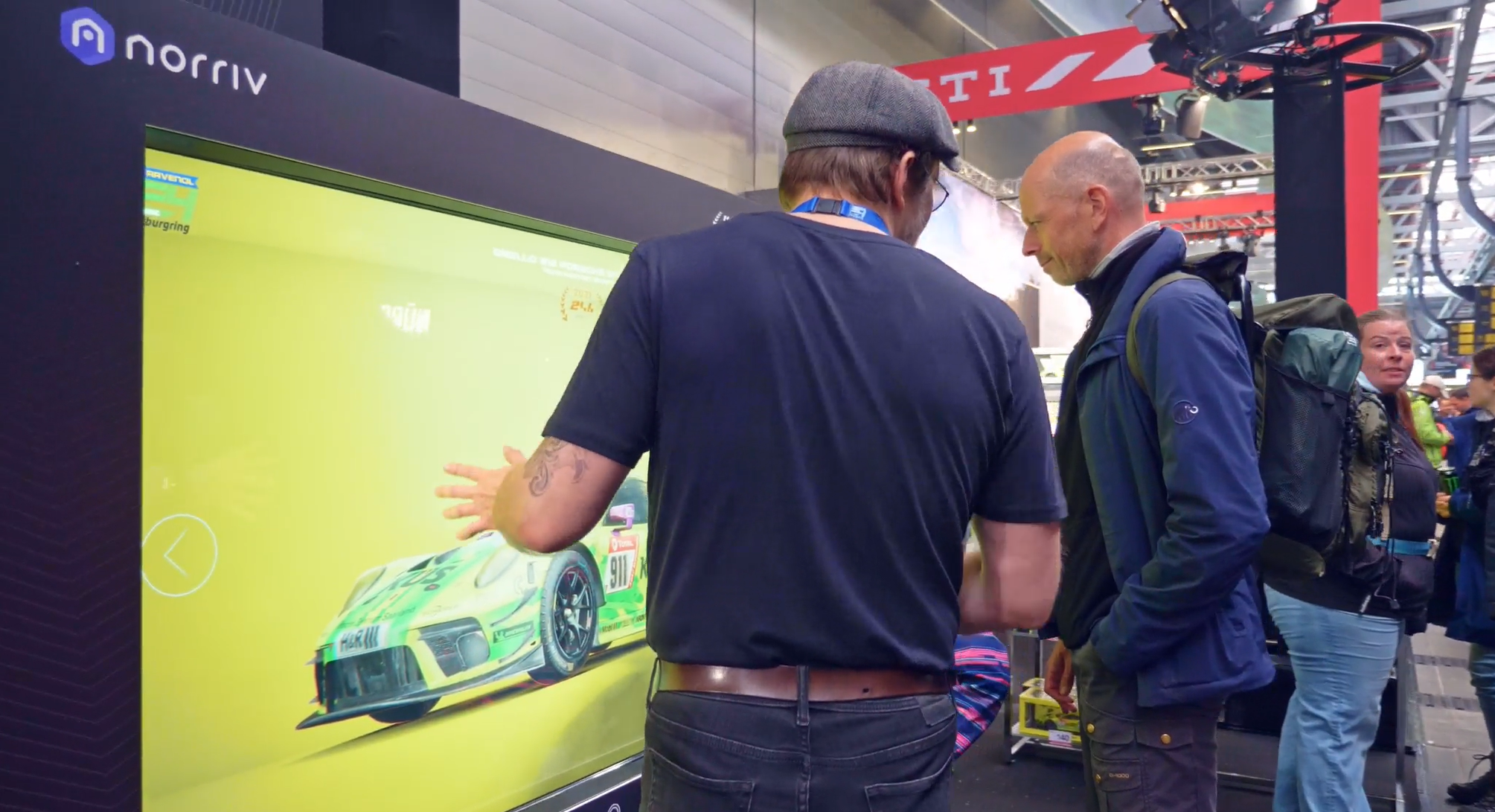 A person wearing a cap interacts with a large touchscreen displaying a green race car while others watch at an indoor event or exhibition booth.