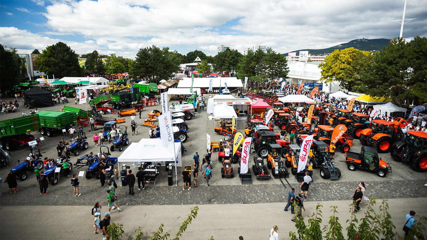 A bustling outdoor exhibition features tractors, farming equipment, vendor tents, and crowds strolling between displays on a cloudy day.