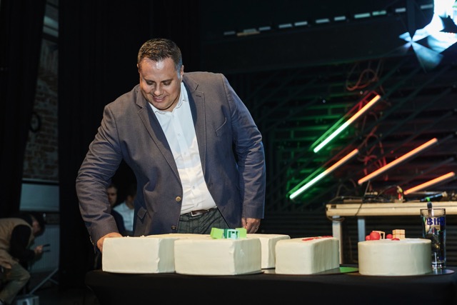 A man in a blue blazer and white shirt smiles as he prepares to cut a large cake on a table. Colorful lights and equipment are visible in the background, suggesting a celebratory event.