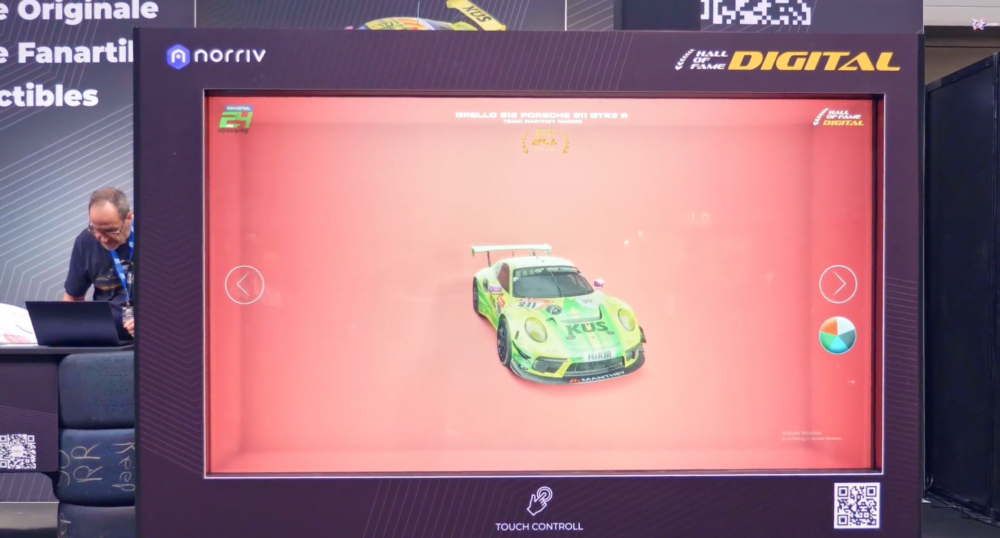 A large digital screen displays a 3D model of a green and pink Porsche race car. A man stands nearby, looking down at a laptop. The screen shows the words “DIGITAL” and “TOUCH CONTROL” at the top and bottom, respectively.