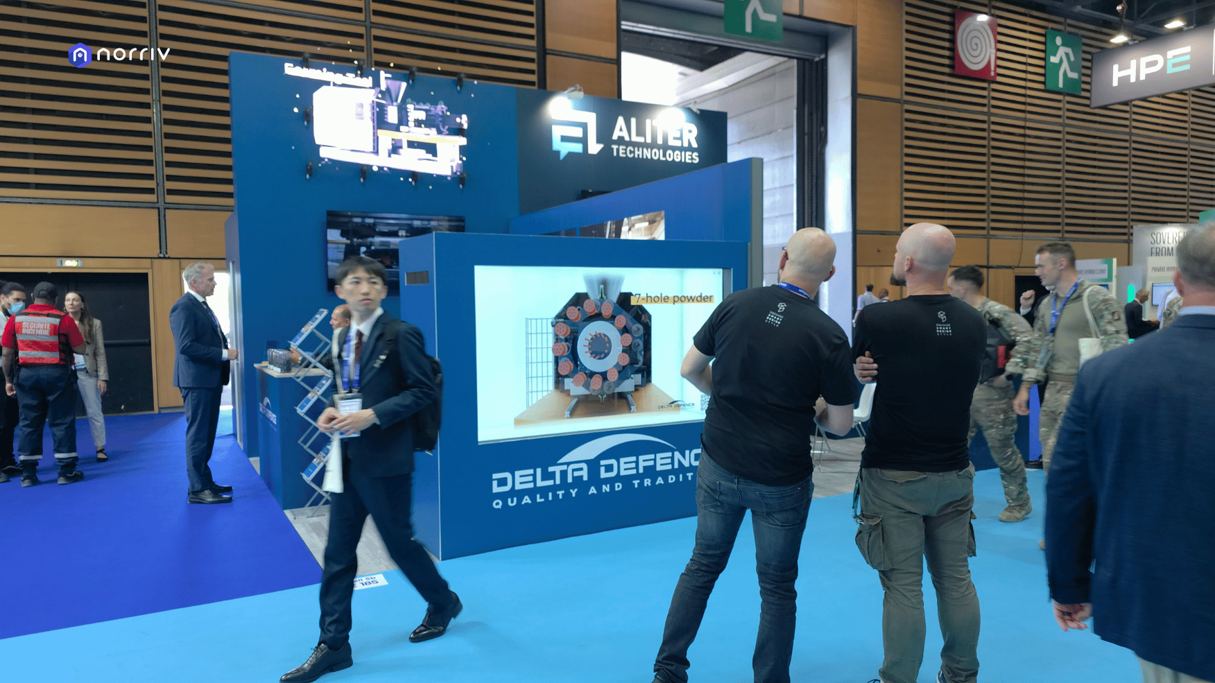 Attendees beside DELTA-DEFENCE's Hologram Box showing a circular machine component, with elevated holographic product visuals behind them.