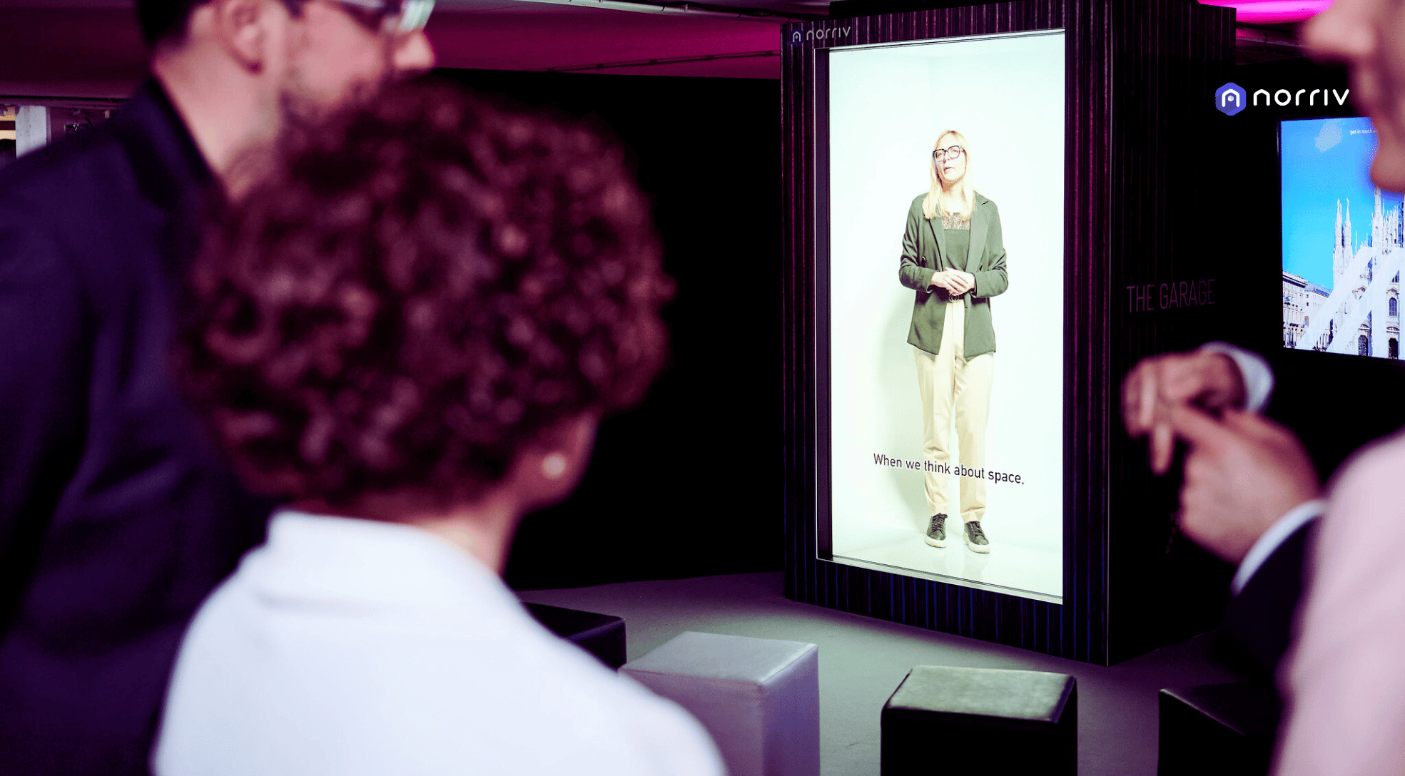 People observe a large digital display featuring a woman standing and talking, with the words Within the finish about space. The display is branded with norriv in the top right corner. The setting appears to be an indoor event or exhibition.