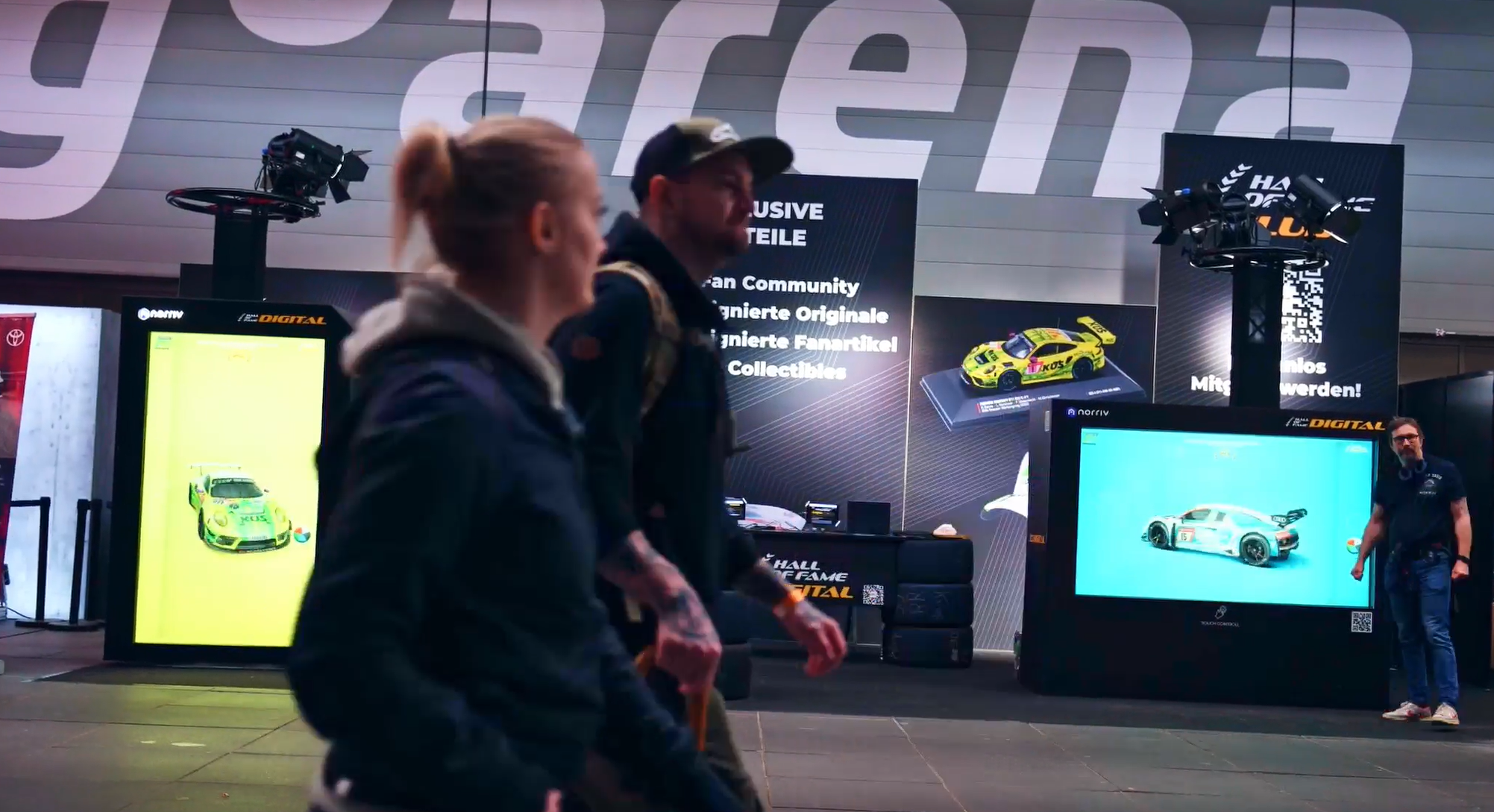 Two people walk past a racing-themed exhibit with car models, display screens, and large signage reading arena in the background. The area is brightly lit, showcasing collectible items and digital art.
