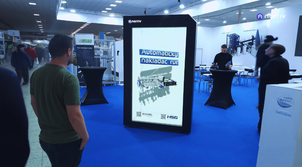 A man stands in front of a large vertical digital display at an indoor trade show. The screen shows text in Czech and technical images. Other attendees and exhibition tables are visible in the background.