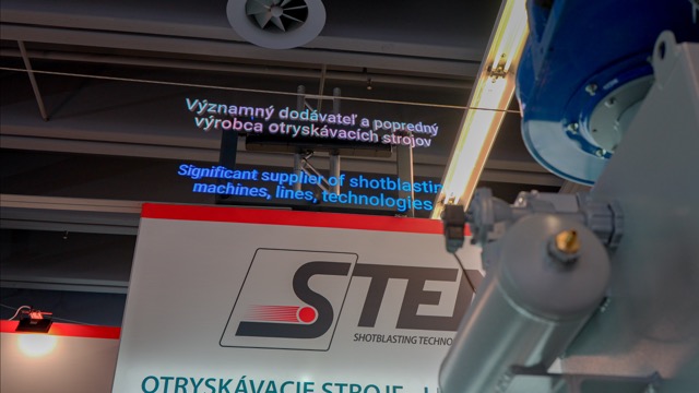 A trade show booth displays the STEE logo with the slogan Significant supplier of shotblasting machines, lines, technologies projected in English and Slovak above the booth. Industrial equipment is partially visible in the foreground.