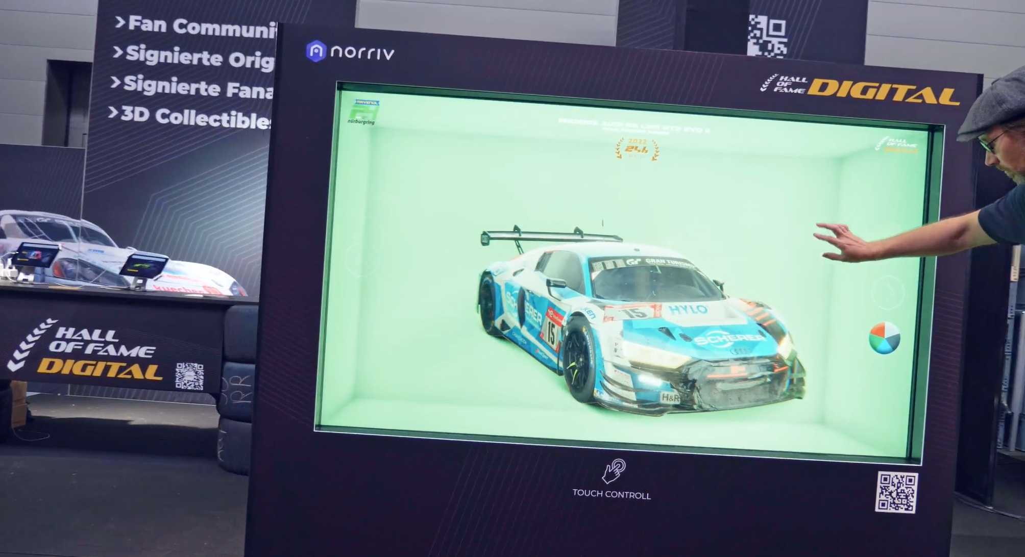 A person interacts with a large digital display showing a 3D model of a blue and white race car. Another race car and Hall of Fame Digital signage are visible in the background.