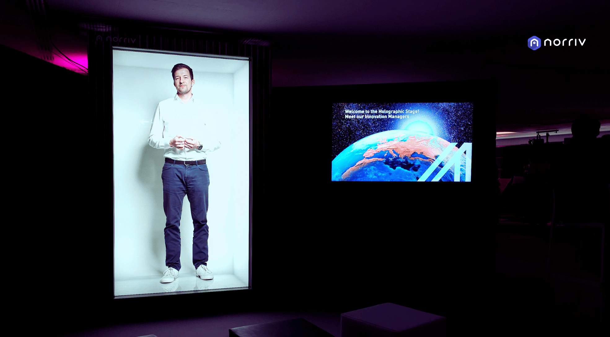 A man stands inside a large, lit rectangular frame, appearing like a hologram. Next to him is a screen displaying an image of Earth from space and text. The scene is indoors with dim lighting.