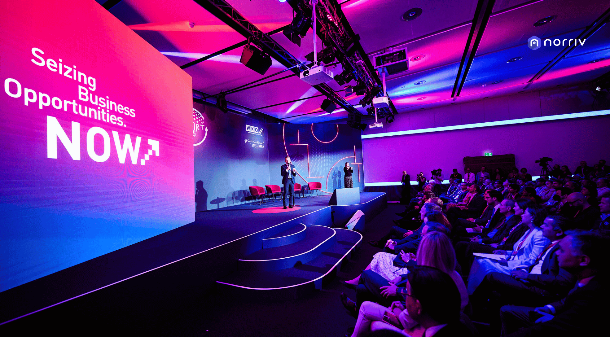 A large conference room with purple and pink lighting, a speaker on stage near a screen displaying “Seizing Business Opportunities. NOW.” and an audience seated, attentively watching the presentation.