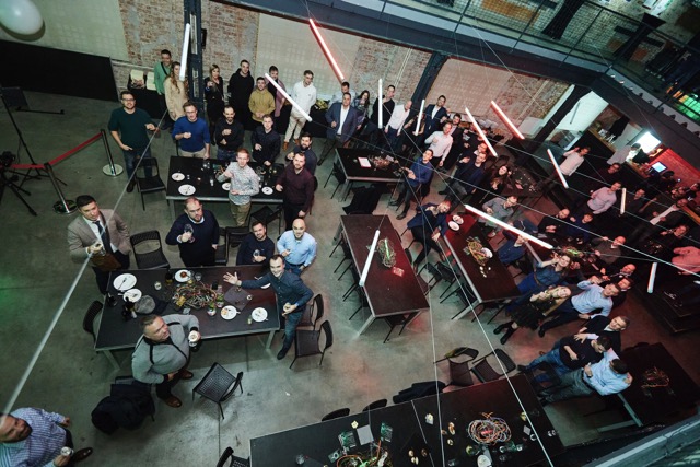 A group of people at an indoor event, standing and sitting around tables with food and drinks, looking up at the camera. The room has exposed brick walls and hanging lights.