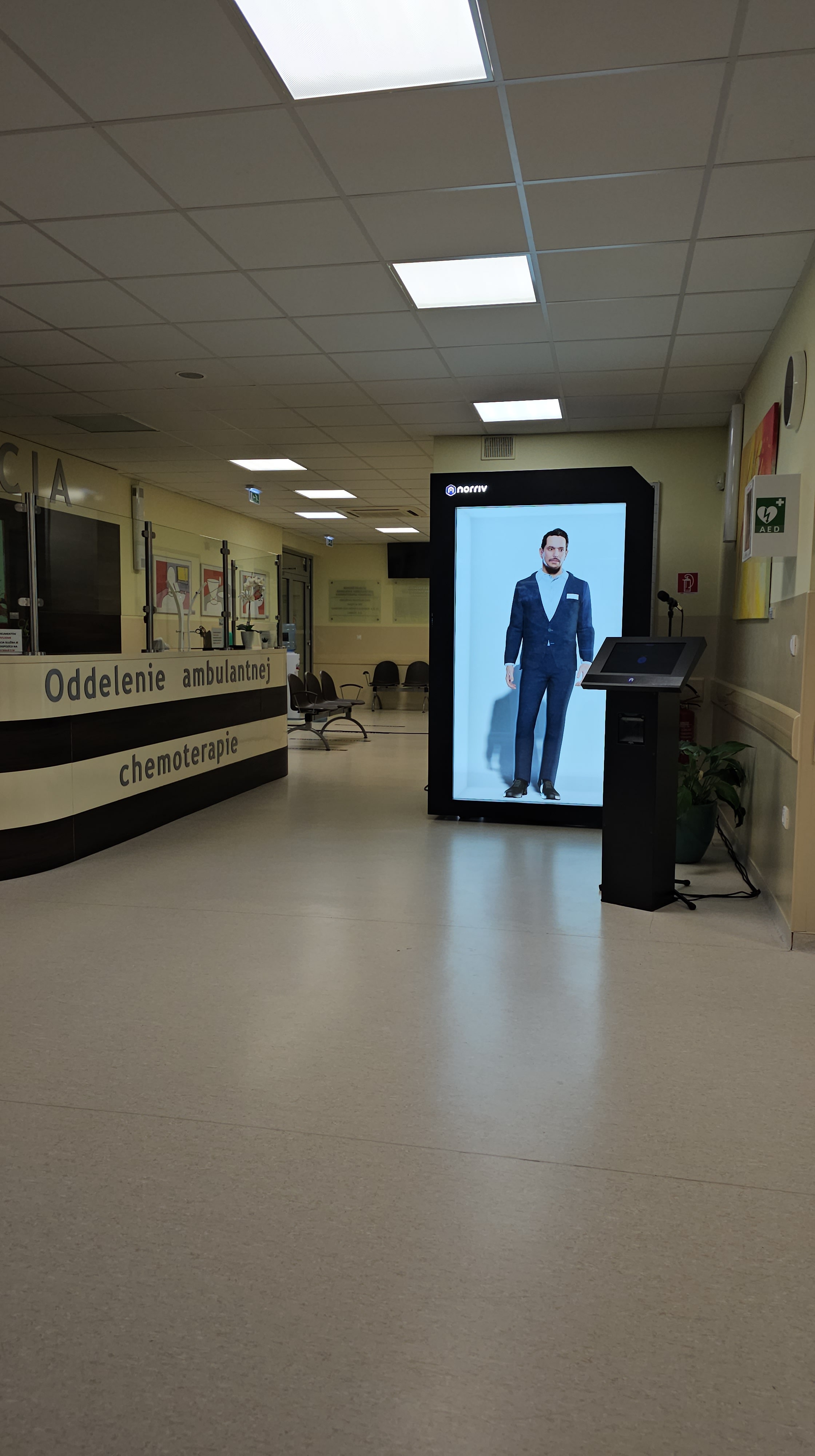 A hospital hallway with empty chairs and a reception desk labeled Oddelenie ambulantnej chemoterapie. A digital screen displays a man in a suit near the entrance. The area is clean and well-lit.