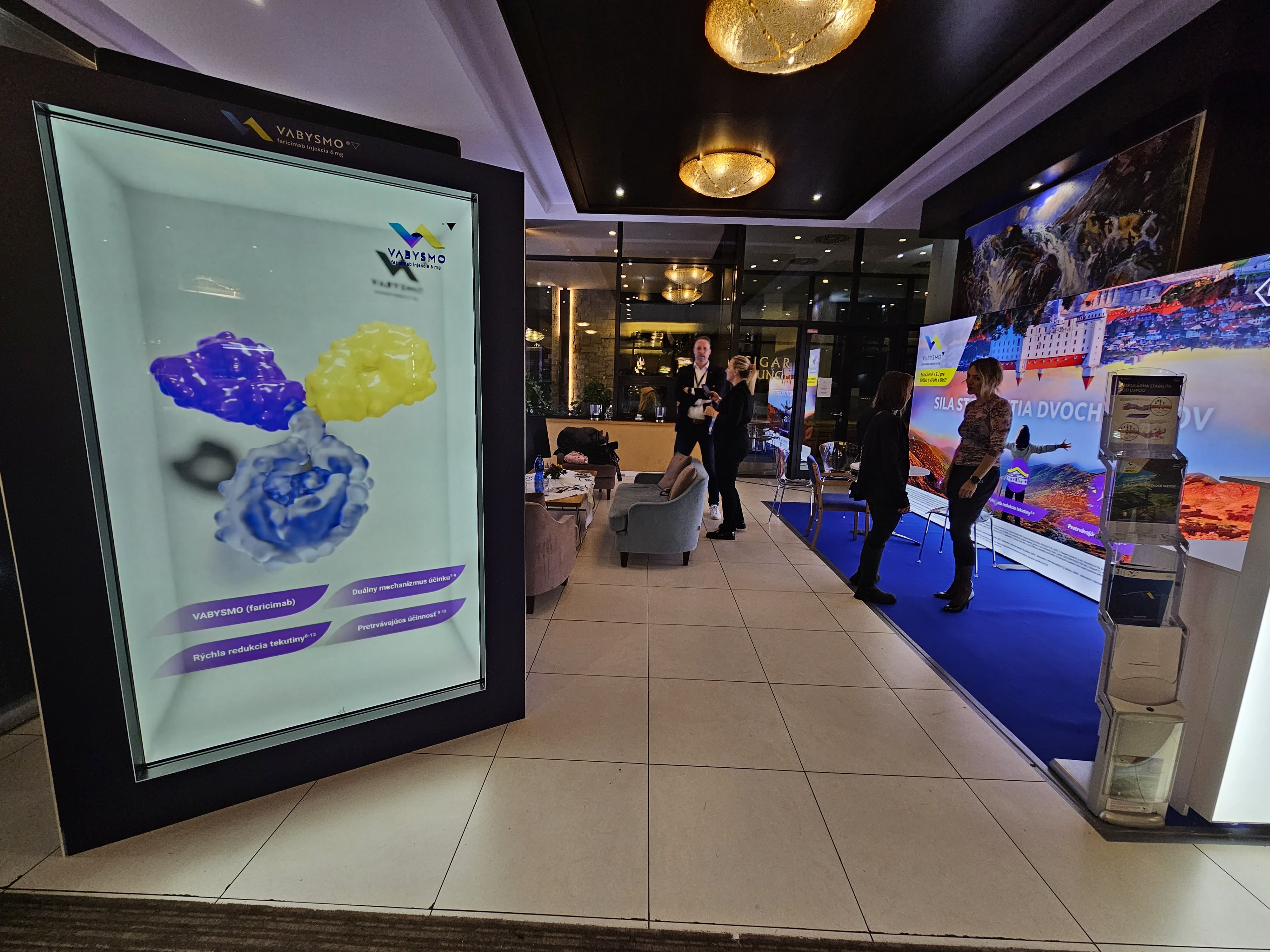 A brightly lit exhibition booth features a digital display showing colorful molecular models, with people interacting nearby and promotional materials visible in the background.