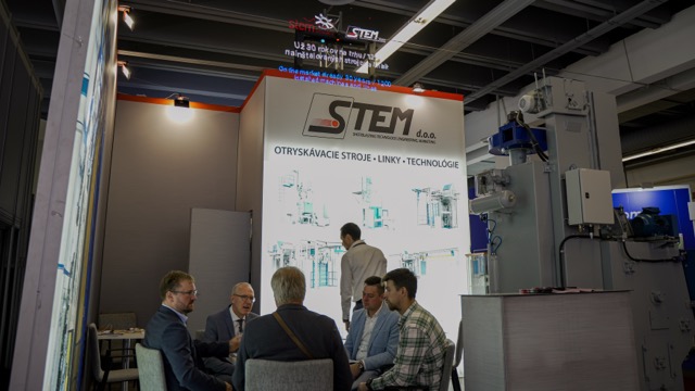 Six people are having a discussion at a trade show booth labeled STEM, with machinery diagrams and equipment displayed in the background. The booth is well-lit and features industrial technology.