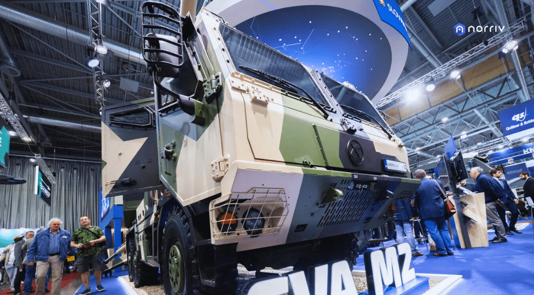 A large, camouflaged military truck is displayed at an indoor exhibition, with people walking around and observing the vehicle. Booths and banners are visible in the background.