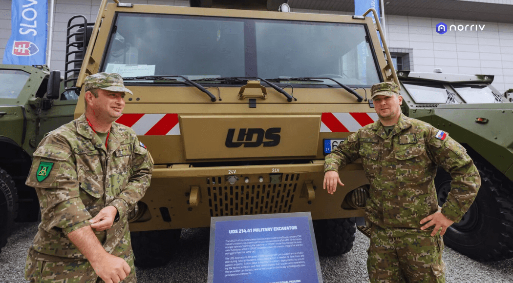 Two soldiers in camouflage uniforms stand in front of a large military vehicle with UDS written on it, displayed outdoors. A sign with information is positioned between them.