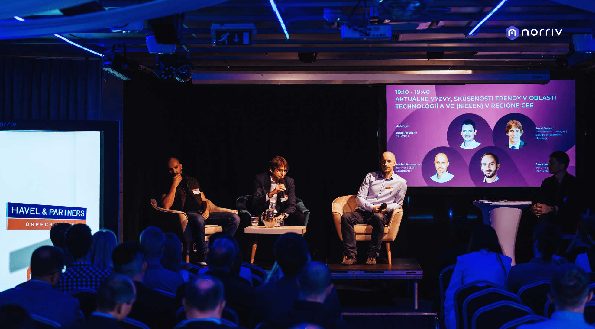 A panel of four speakers sits on a stage in front of an audience at a tech conference, with a large screen behind them displaying their names, photos, and the event topic in Czech. Blue and purple lighting fills the room.
