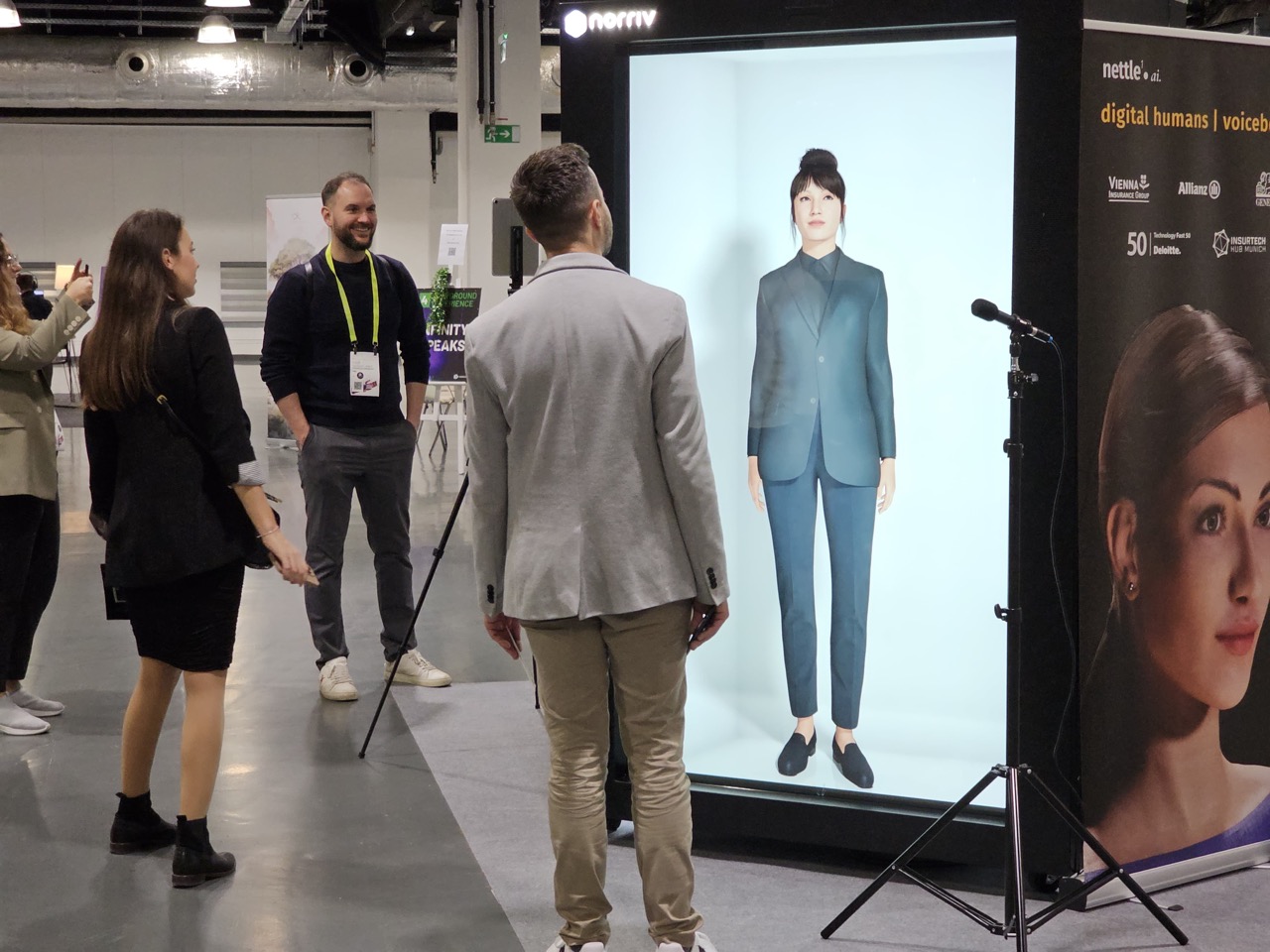 Several people interact with a large digital screen displaying a lifelike female avatar at a tech event. One man stands directly in front of the avatar, while others watch and smile nearby.