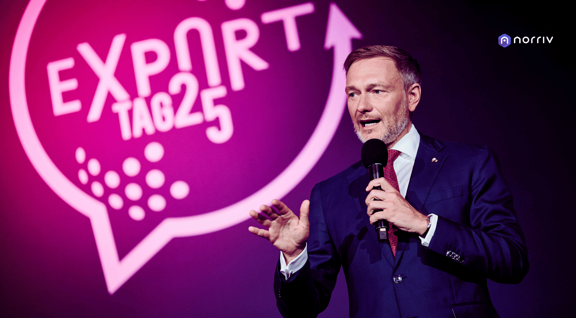 A man in a blue suit speaks into a microphone on stage in front of a large, pink sign that reads EXPORT TAG 25 with an upward arrow and a dotted pattern. The logo norriv appears in the top right corner.