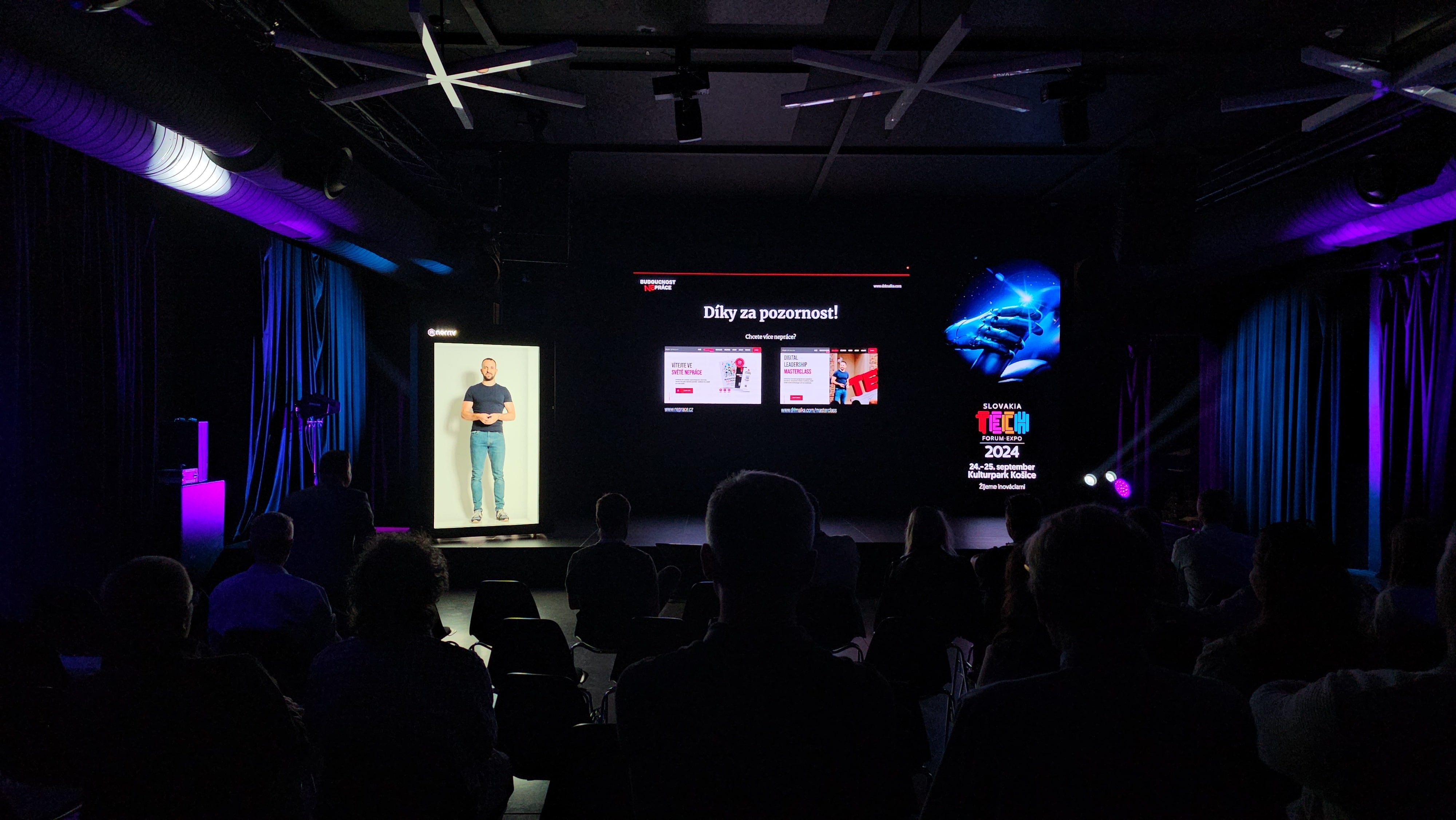 A dark conference room with people seated, watching a presentation on a large screen. The screen shows slides and a presenter; blue and purple stage lighting highlights the space.