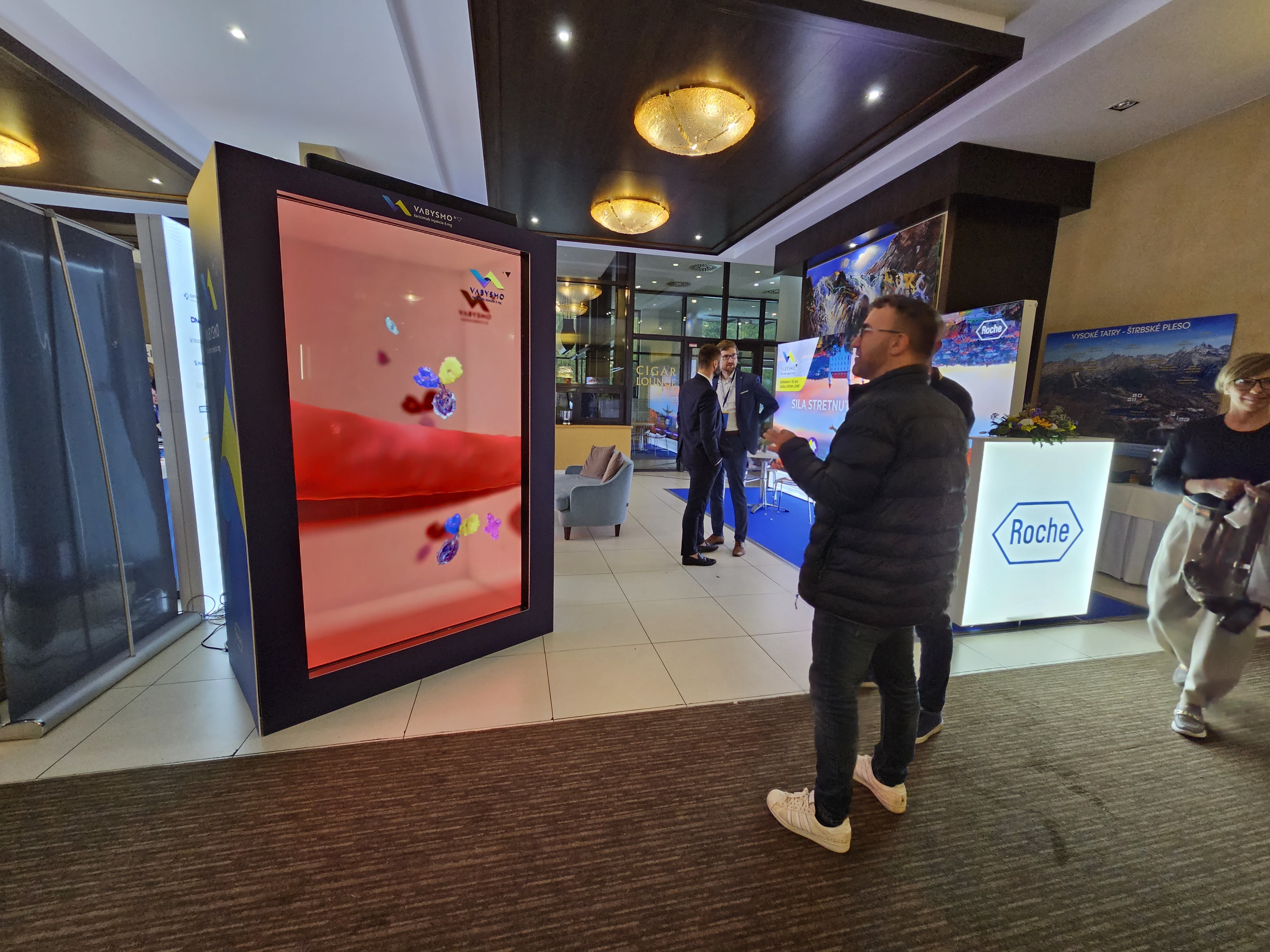 Two people stand in a modern, well-lit lobby near a large vertical digital display showing colorful graphics. A reception desk with the Roche logo and promotional banners are visible in the background.