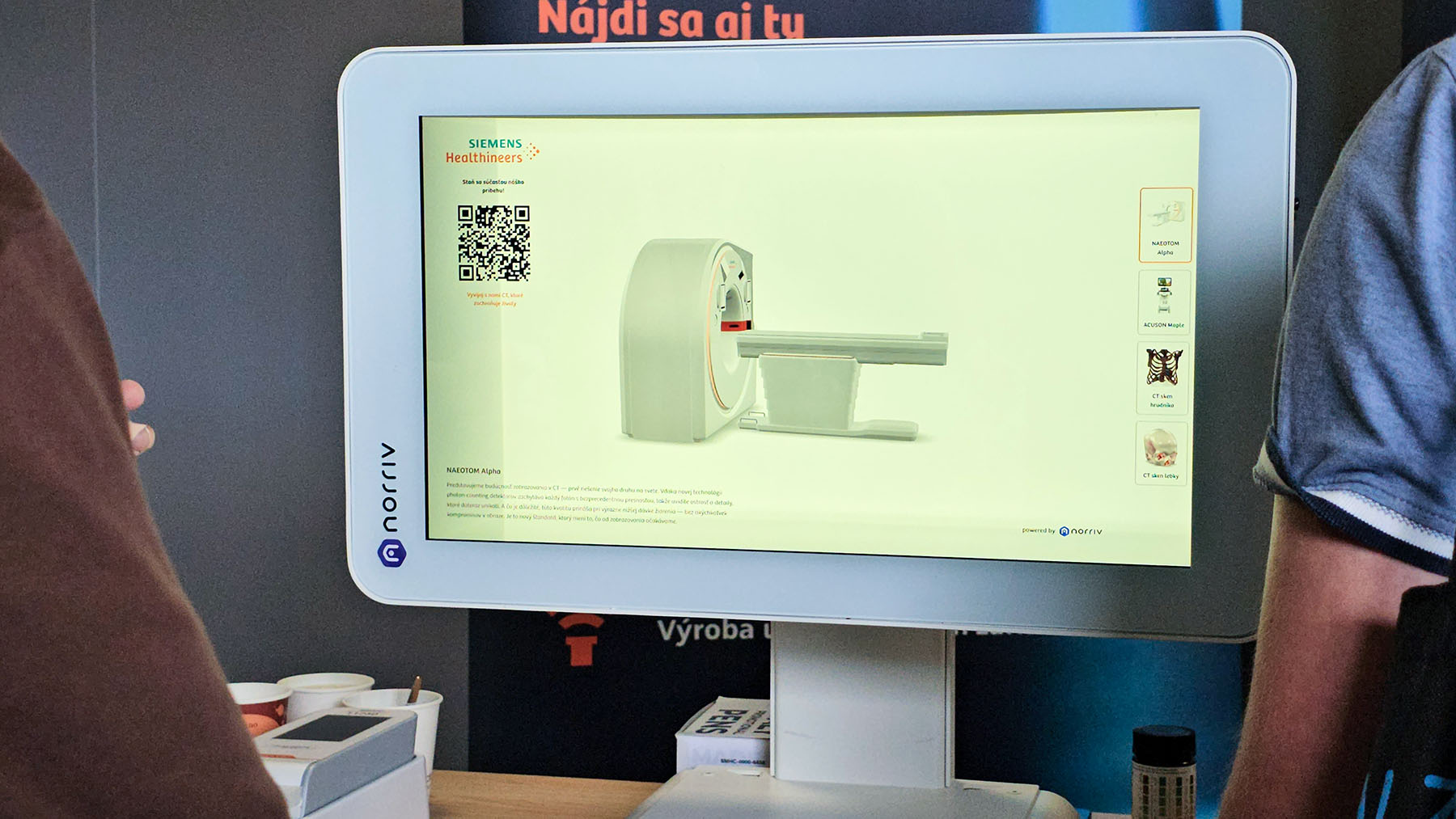 A computer monitor shows a Siemens Healthineers medical imaging device, QR code, and details as two people stand nearby.