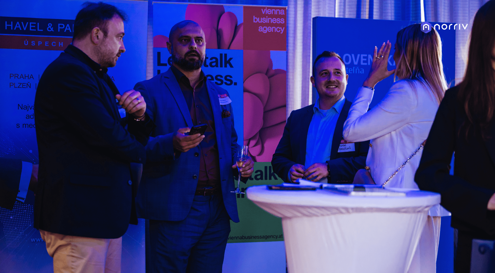 Four people stand and converse at a networking event in front of colorful business banners. Three men wear suits, while a woman in a white jacket gestures with her hand beside a tall round table.