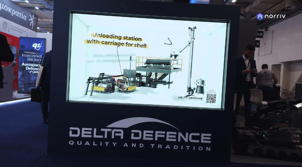 A large display at a trade show features a graphic of an unloading station for shells by Delta Defence, with people and exhibition booths visible in the background. The screen reads “Unloading station with carriage for shell.”.