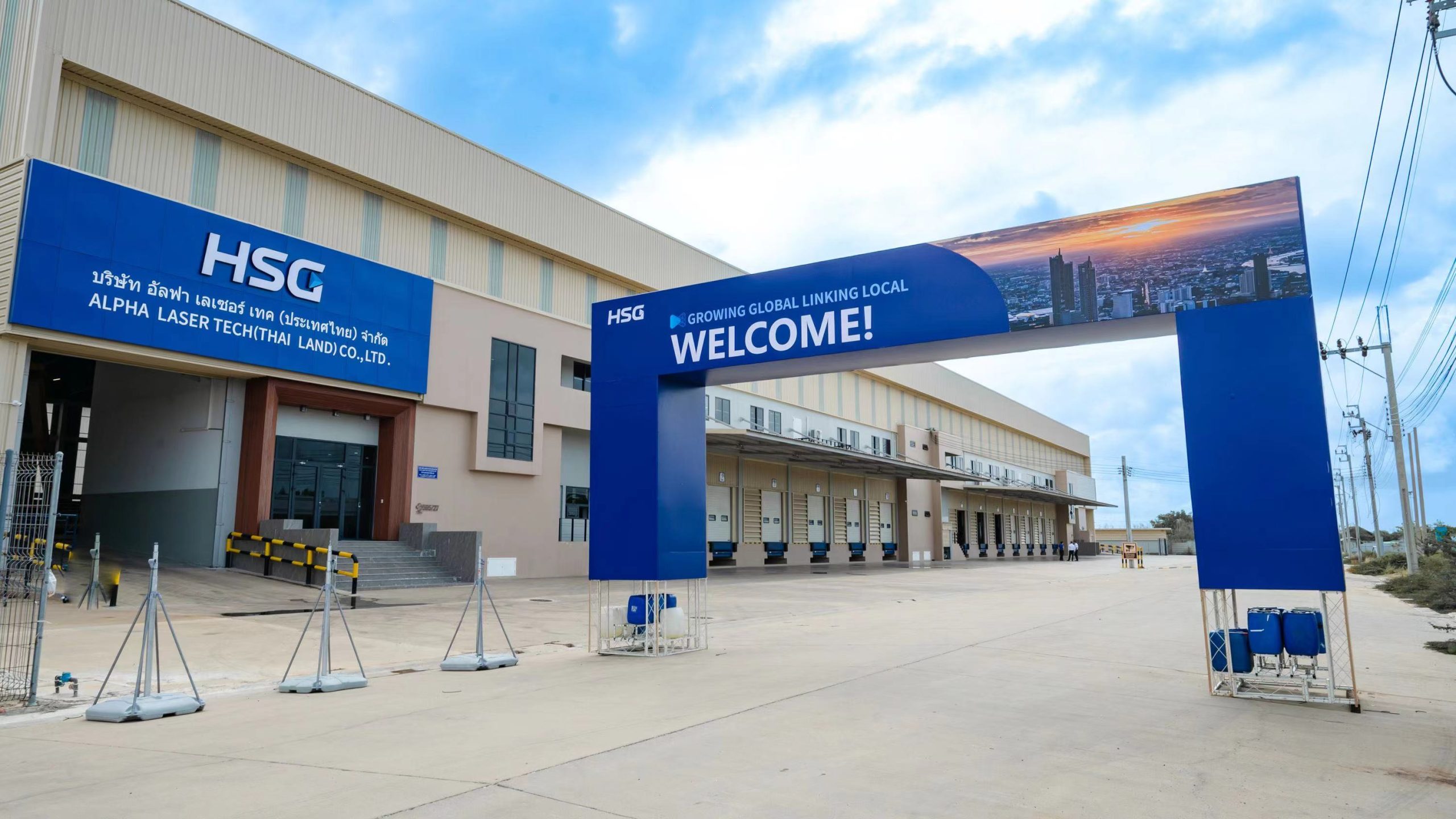 A large industrial facility with a blue HSG sign and a welcoming archway that reads GROWING GLOBAL LINKING LOCAL WELCOME! in front of a loading dock area under a partly cloudy sky.