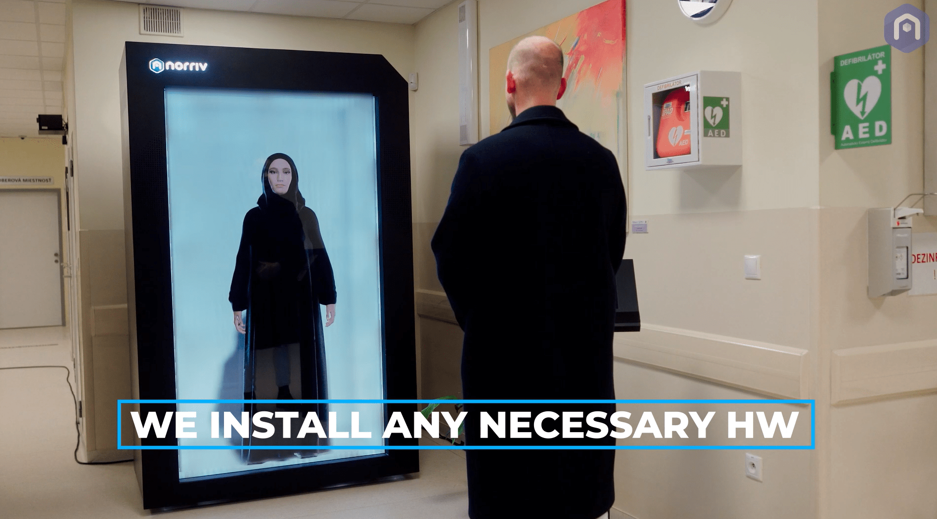 A person in a coat stands in front of a large digital screen displaying a woman in black clothing. Text on the screen reads, WE INSTALL ANY NECESSARY HW. The setting appears to be a modern indoor facility.