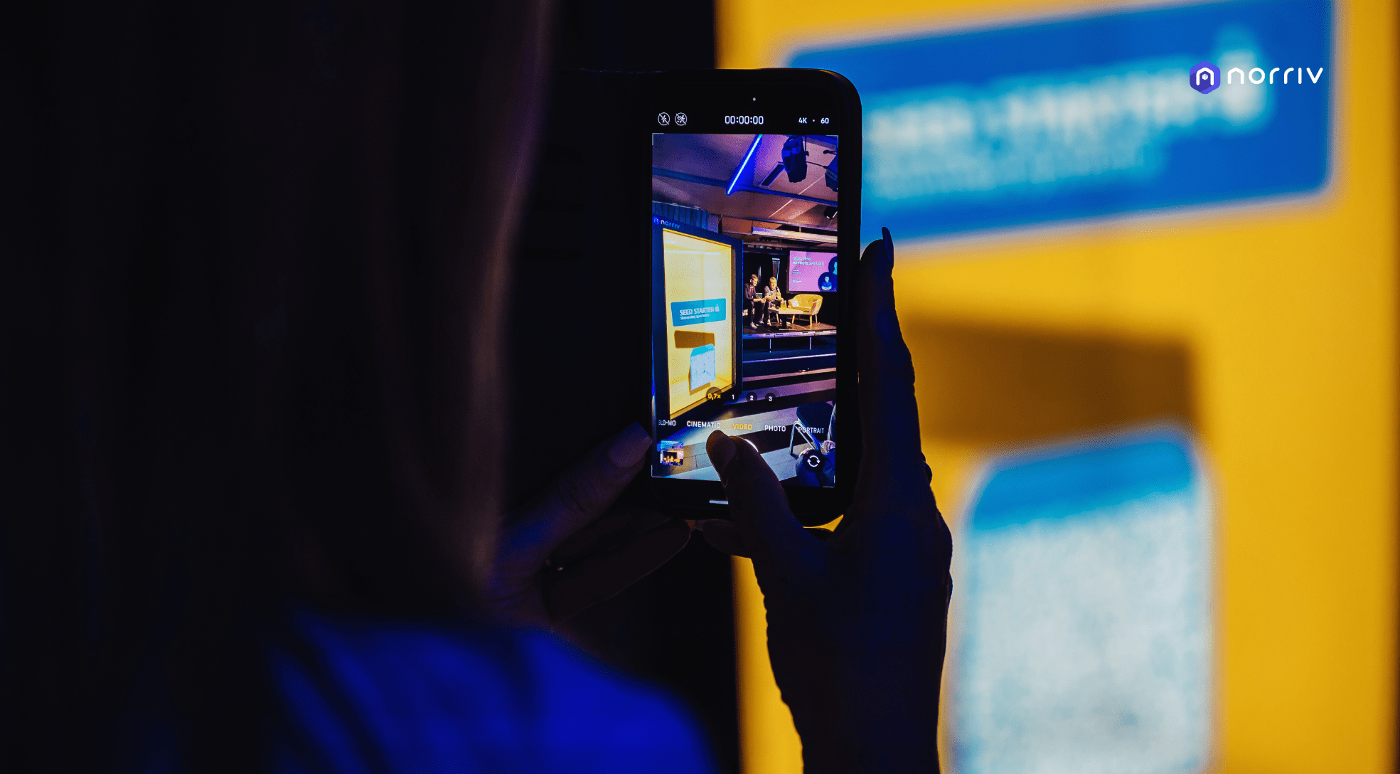 A person takes a photo of a brightly lit screen or display with their smartphone at an event. The scene is illuminated in blue and yellow tones, and a logo reading norriv is visible in the top right corner.