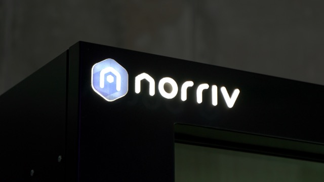 Illuminated norriv logo with a blue hexagonal icon appears on the upper corner of a dark, modern structure against a blurred gray background.