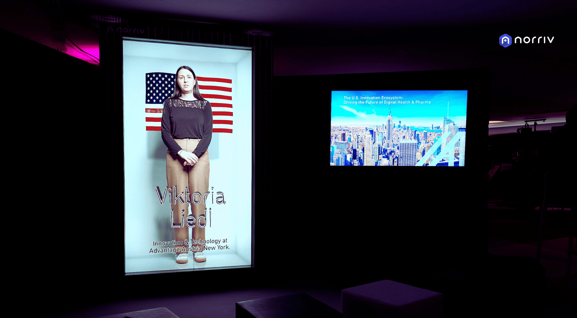 A digital display shows a woman standing in front of an American flag with text reading Viktoria Liedl, Innovation & Technology at Advantage Austria, New York. Another screen shows a city skyline and the words The U.S. Innovation Ecosystem.