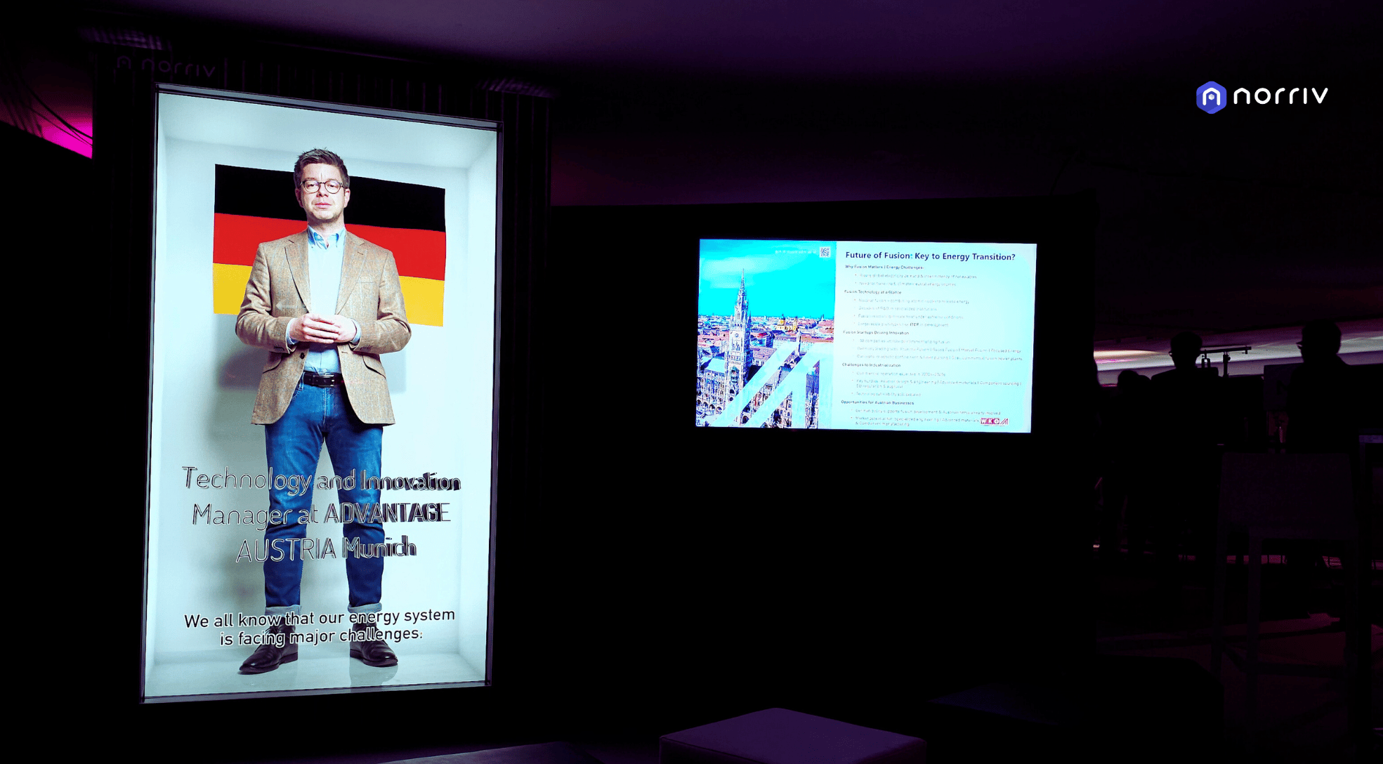 A man stands on a stage next to a large screen showing his image, with the German flag and text about technology, innovation, and energy challenges. Another screen displays information and a cityscape photo.