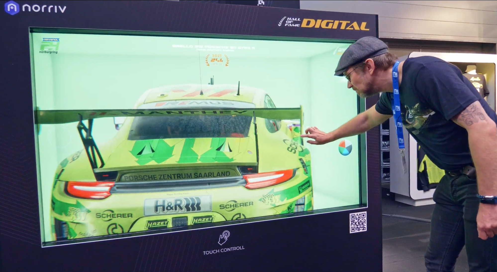 A man touches a digital display showing the rear view of a bright green Porsche race car. The interactive screen features branding, a QR code, and touch control icons, with exhibition signage visible above.