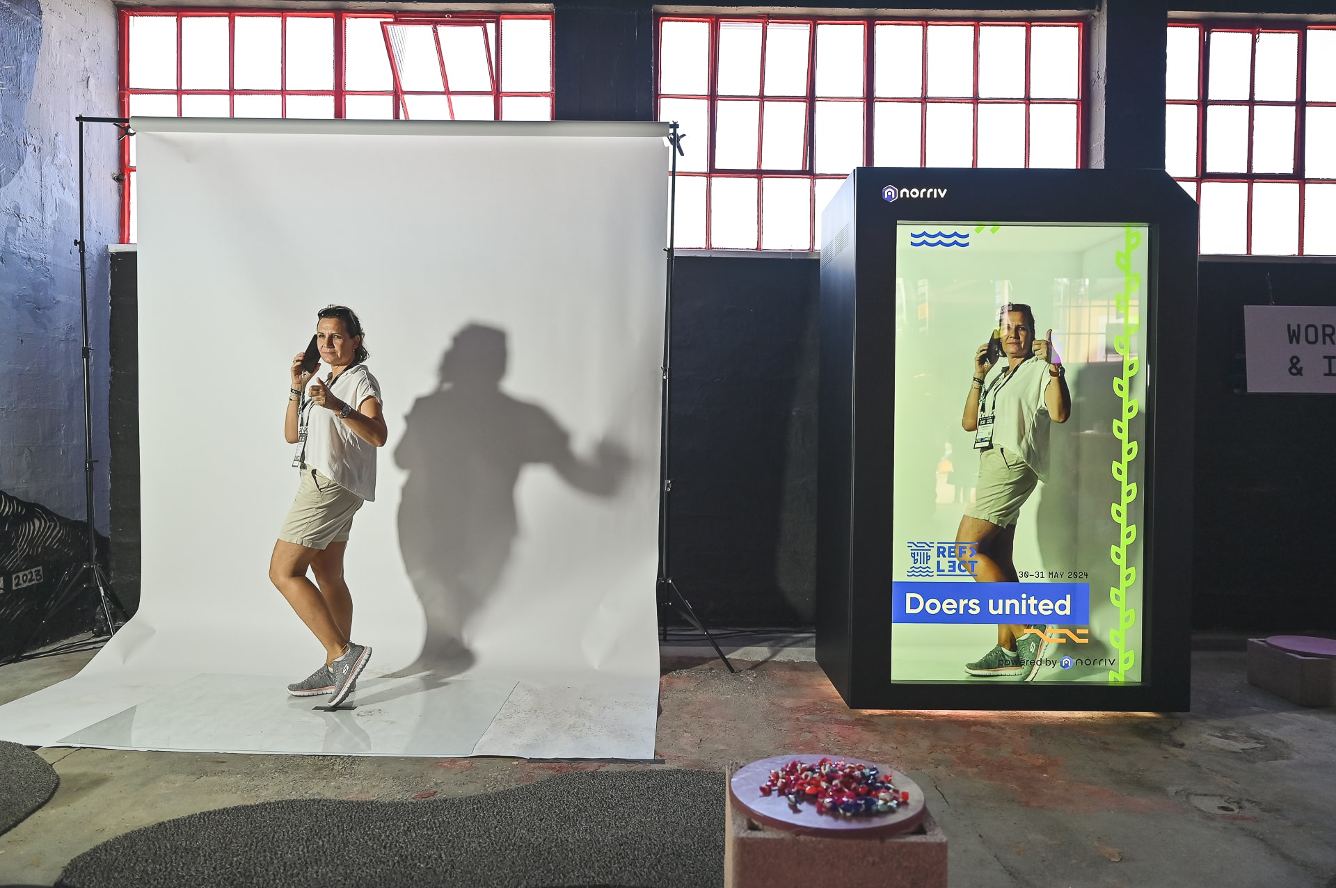 A person poses on a small backdrop for a photo, holding up two finger guns. Next to them, a digital screen displays the same image with decorative graphics and the text Doers united. Red light filters through windows above.