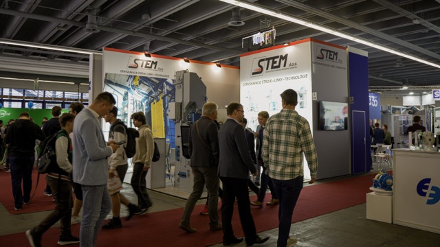 People walk through a trade show with booths and displays under fluorescent lighting. The prominent booth in the center has the sign STEM and features technical equipment and informational posters.